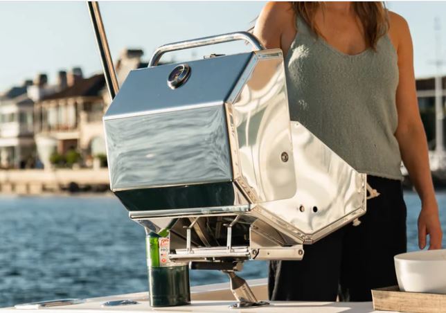 A metallic barbecue grill mounted on a boat railing, with a partial view of a person on a sunny day.